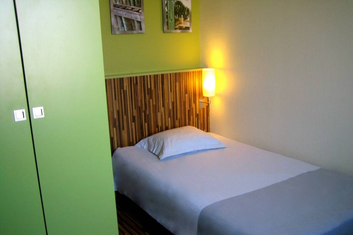 A single bed with a wooden headboard, a bedside lamp, and a green wall with two small pictures.
