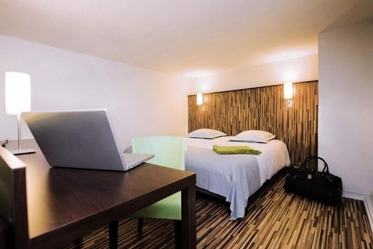 A hotel room with a wooden floor, a bed with white bedding, a desk with a laptop, a green chair, and a black bag.