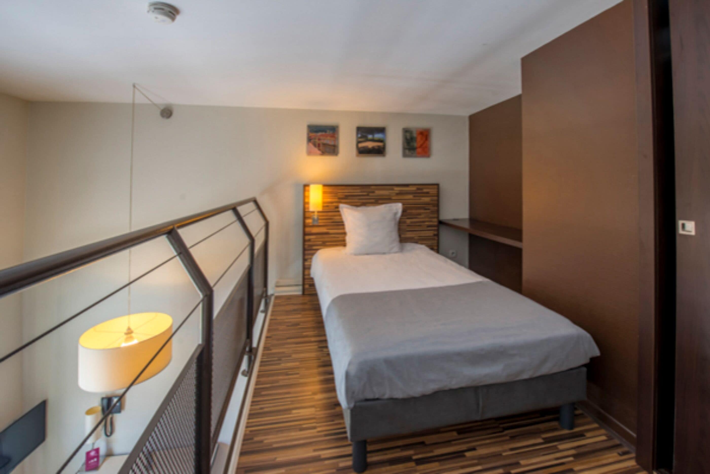 Una camera d'albergo stretta con pavimento in legno, un letto con testiera grigia, un comodino con lampada e una mensola a muro.
