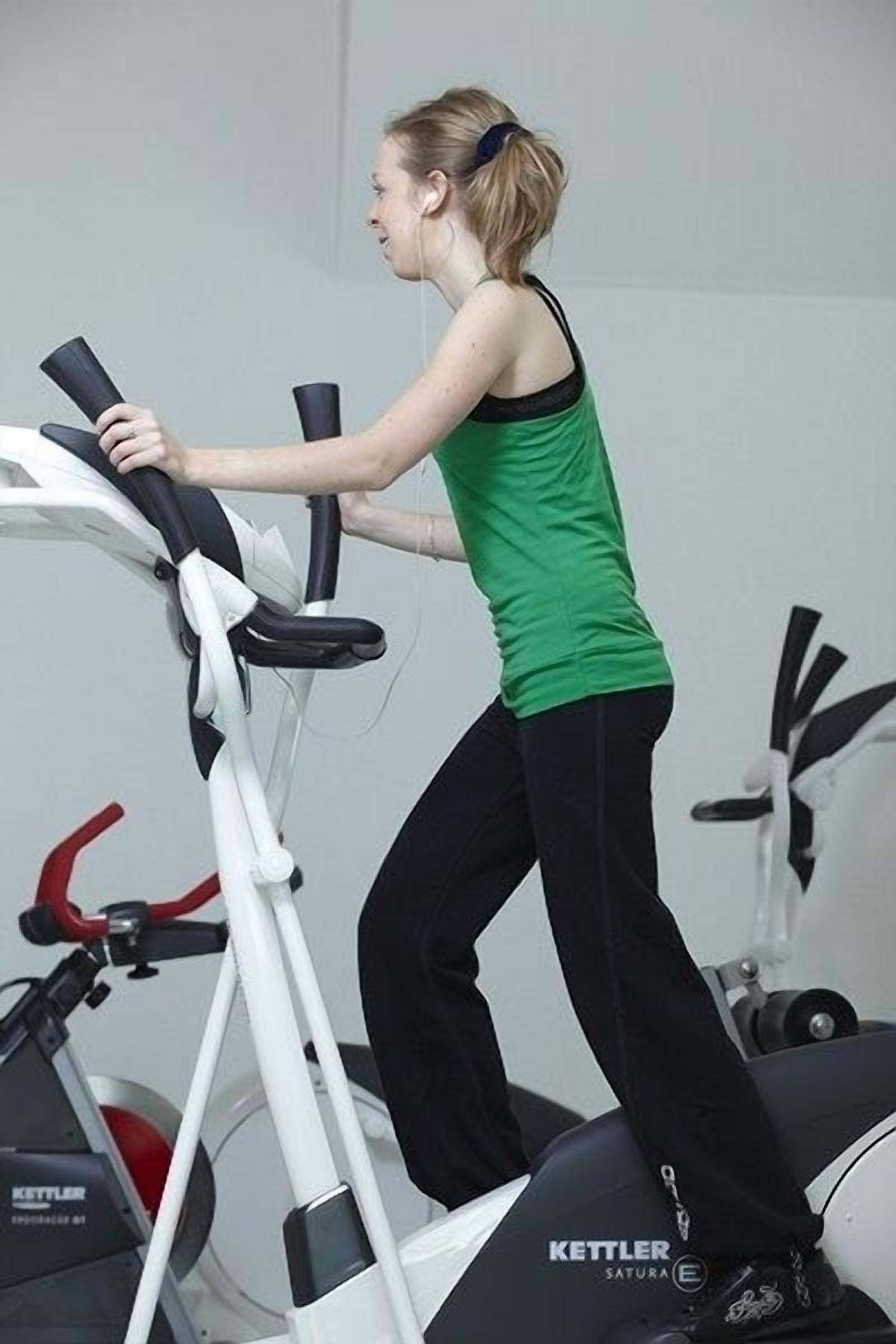 Une personne fait du vélo d’appartement dans une salle de sport, avec d’autres appareils d’exercice en arrière-plan.