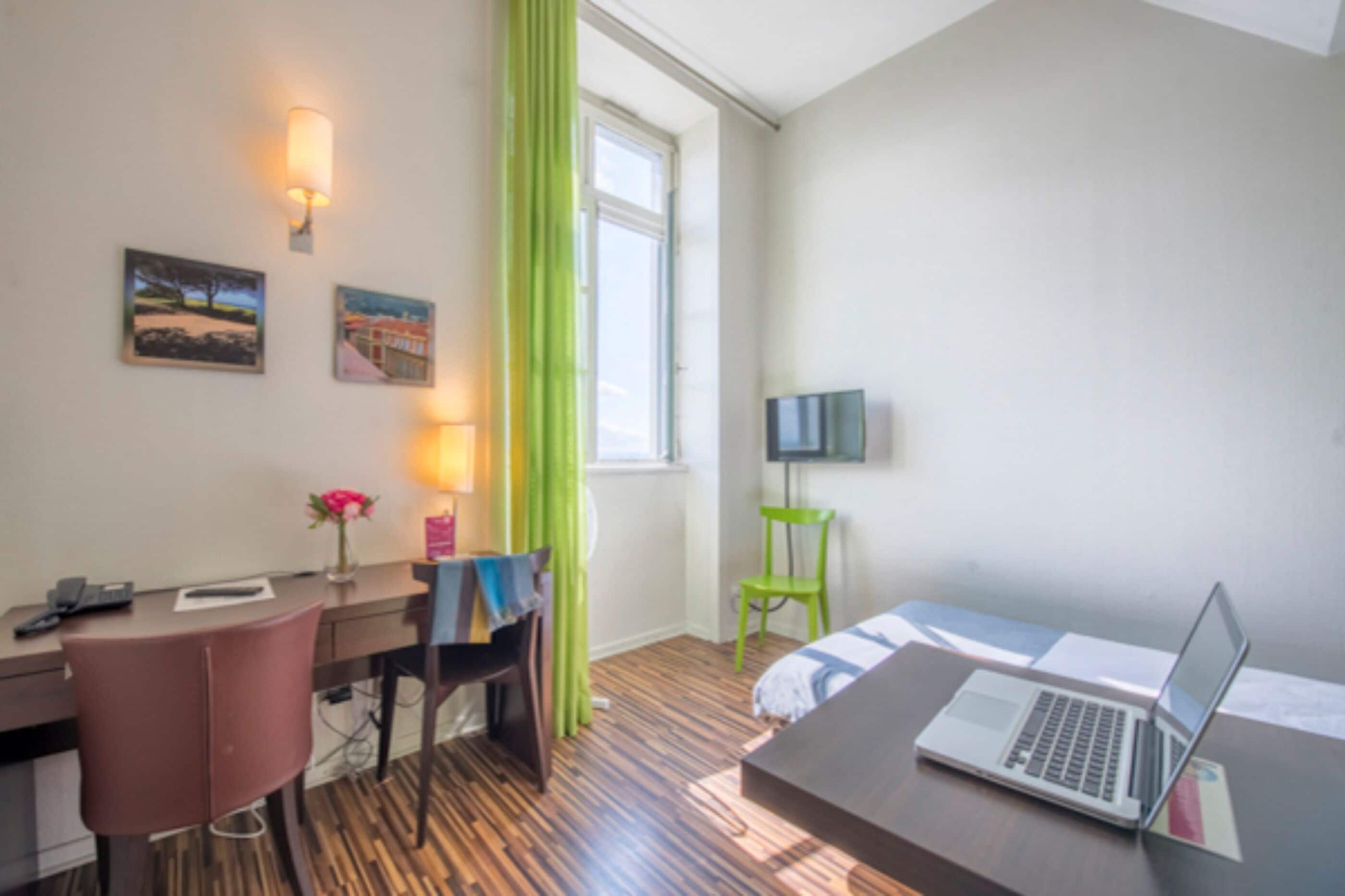 A hotel room with a wooden floor, a desk with a chair, a bed, a TV on the wall, and a window with curtains.