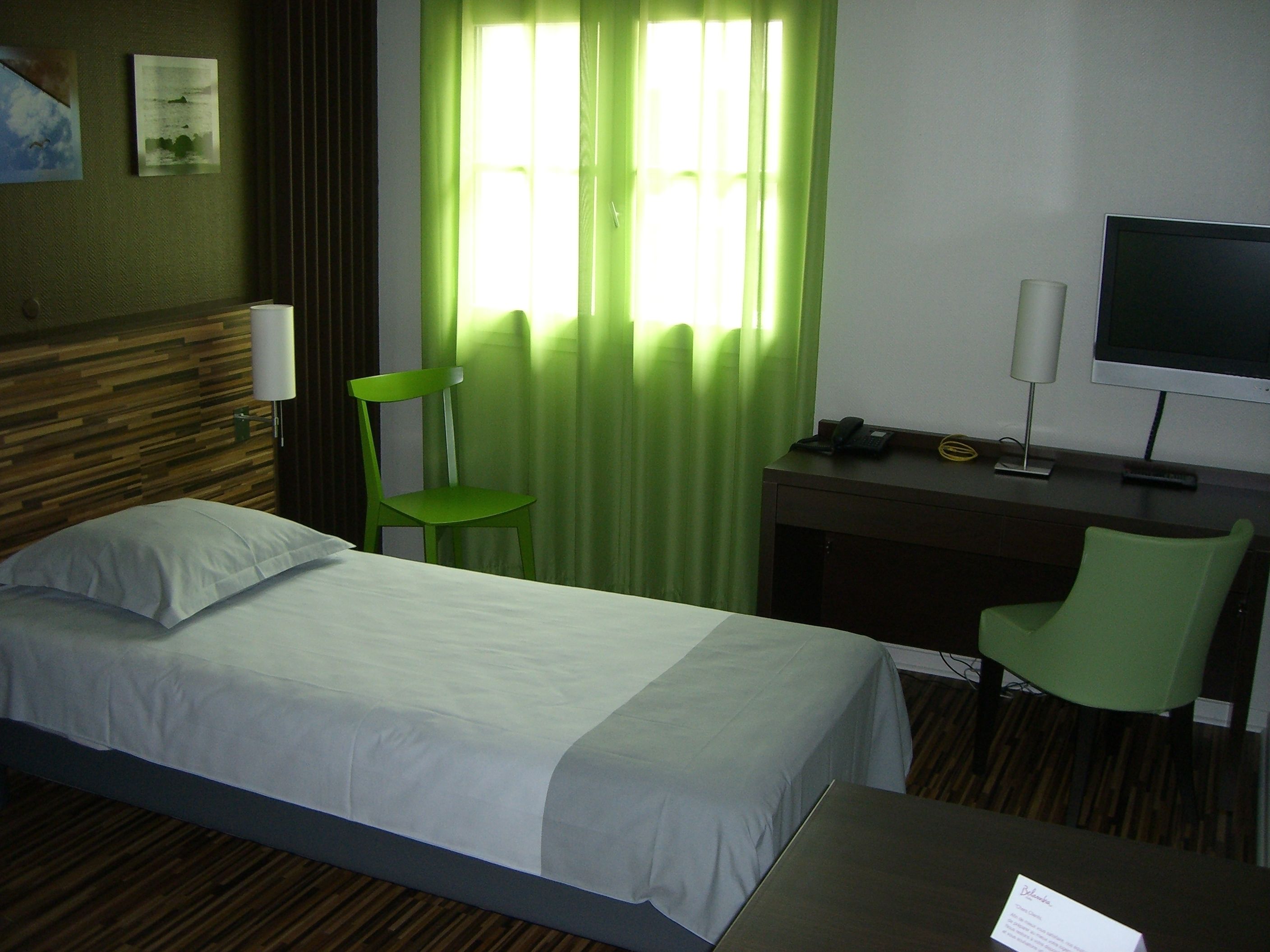 Habitación de hotel con cama, escritorio con silla, televisor y ventana con cortinas verdes.