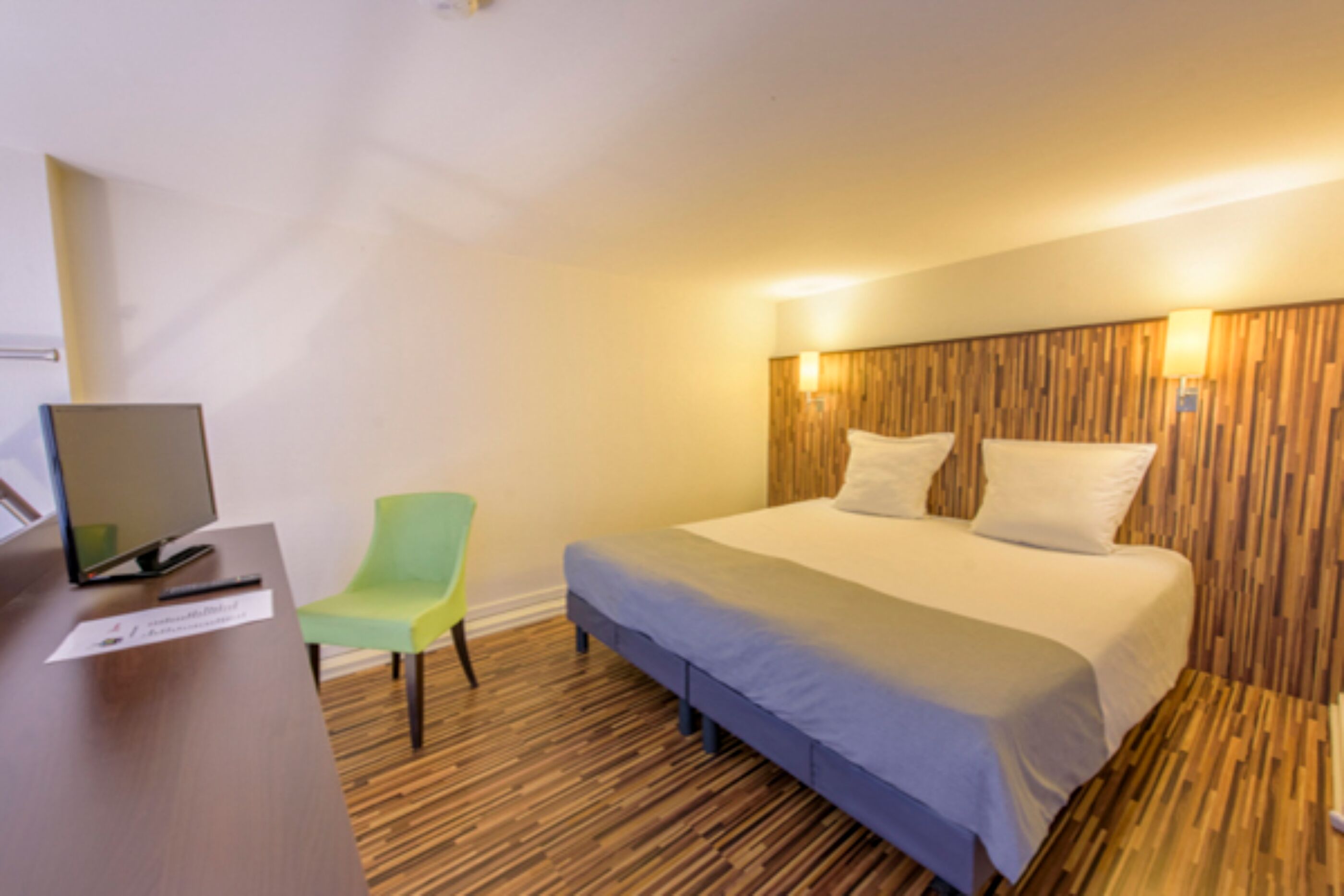 A hotel room with a wooden headboard, a bed with white linens, a desk with a computer, and a green chair.