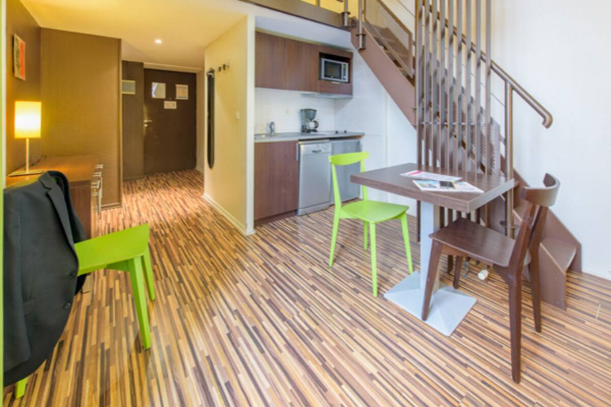 A hotel room with a wooden parquet floor, a dining table with two chairs, a microwave, a small kitchenette, and a staircase.