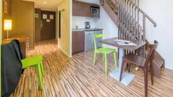 A hotel room with a wooden parquet floor, a dining table with two chairs, a microwave, a small kitchenette, and a staircase.