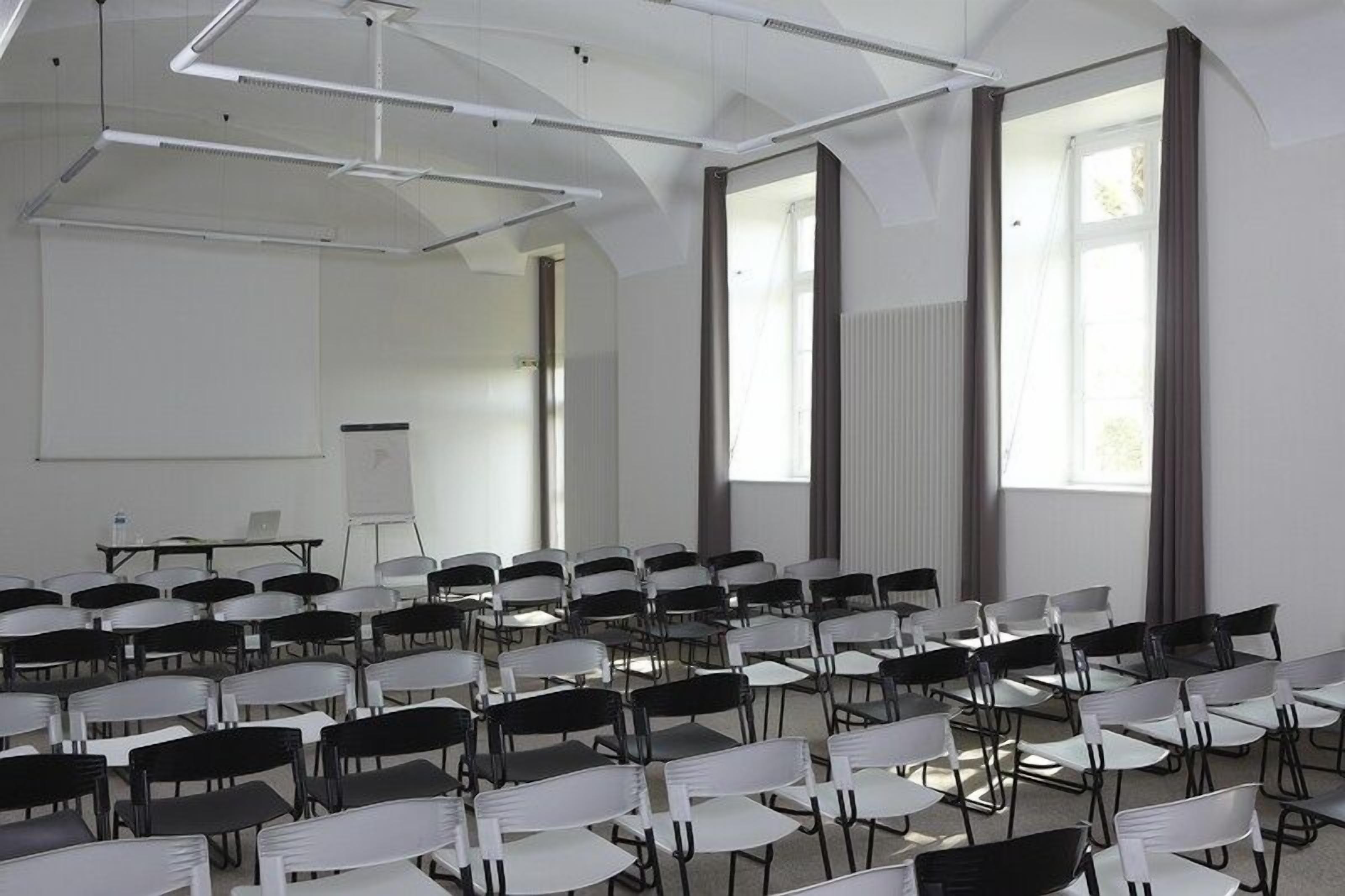 Una sala de conferències amb files de cadires i taules, finestres grans i una pissarra.