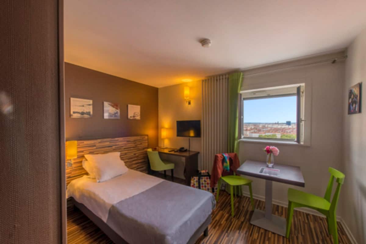 A hotel room with a bed, desk, two chairs, a small table with a vase, and a window with a view.