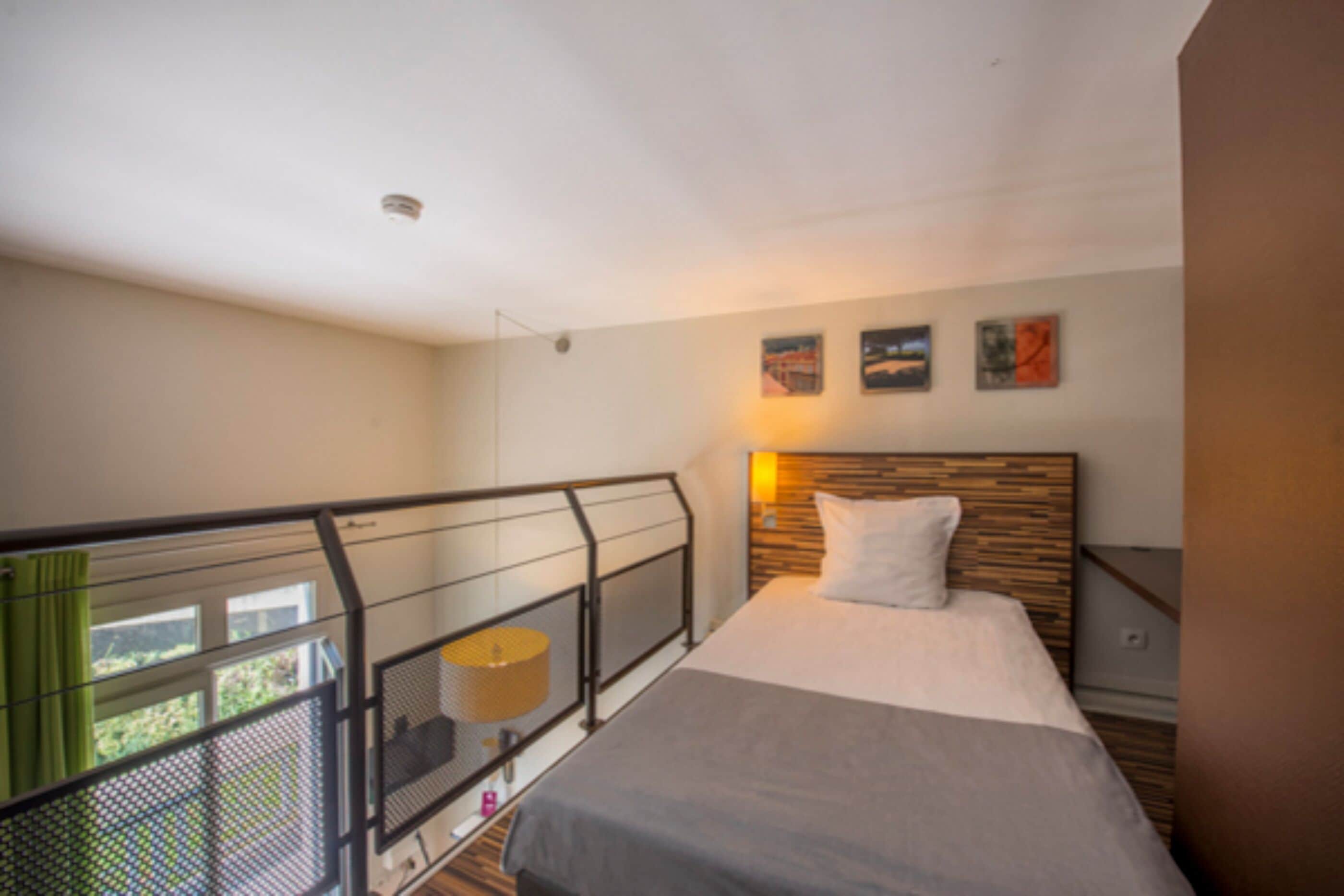 A bedroom with a bed, a nightstand, a lamp, and a view of greenery outside.