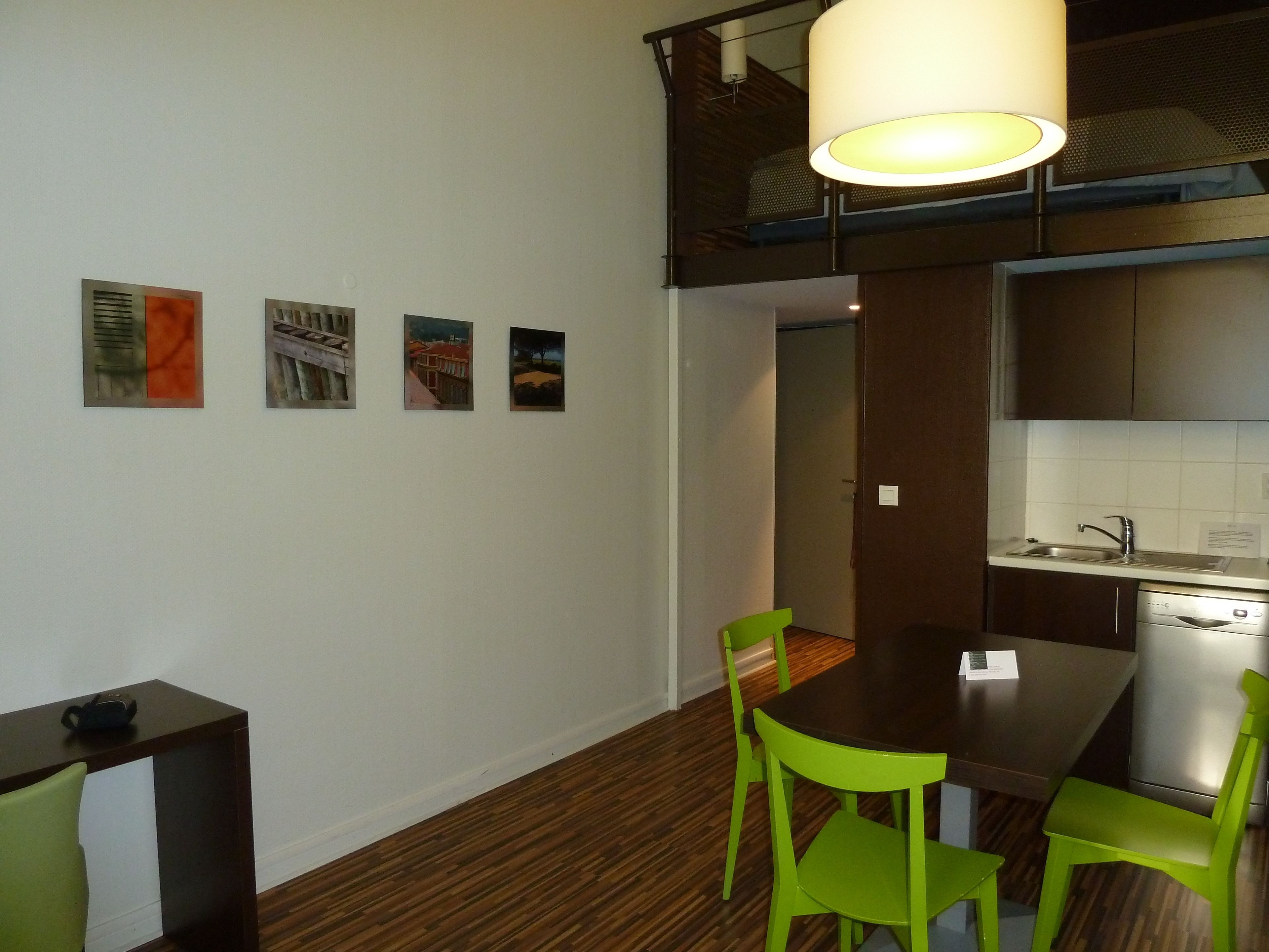 A modern kitchen with a dining area, featuring a wooden floor, green chairs, a dark wooden table, and a hanging light fixture.