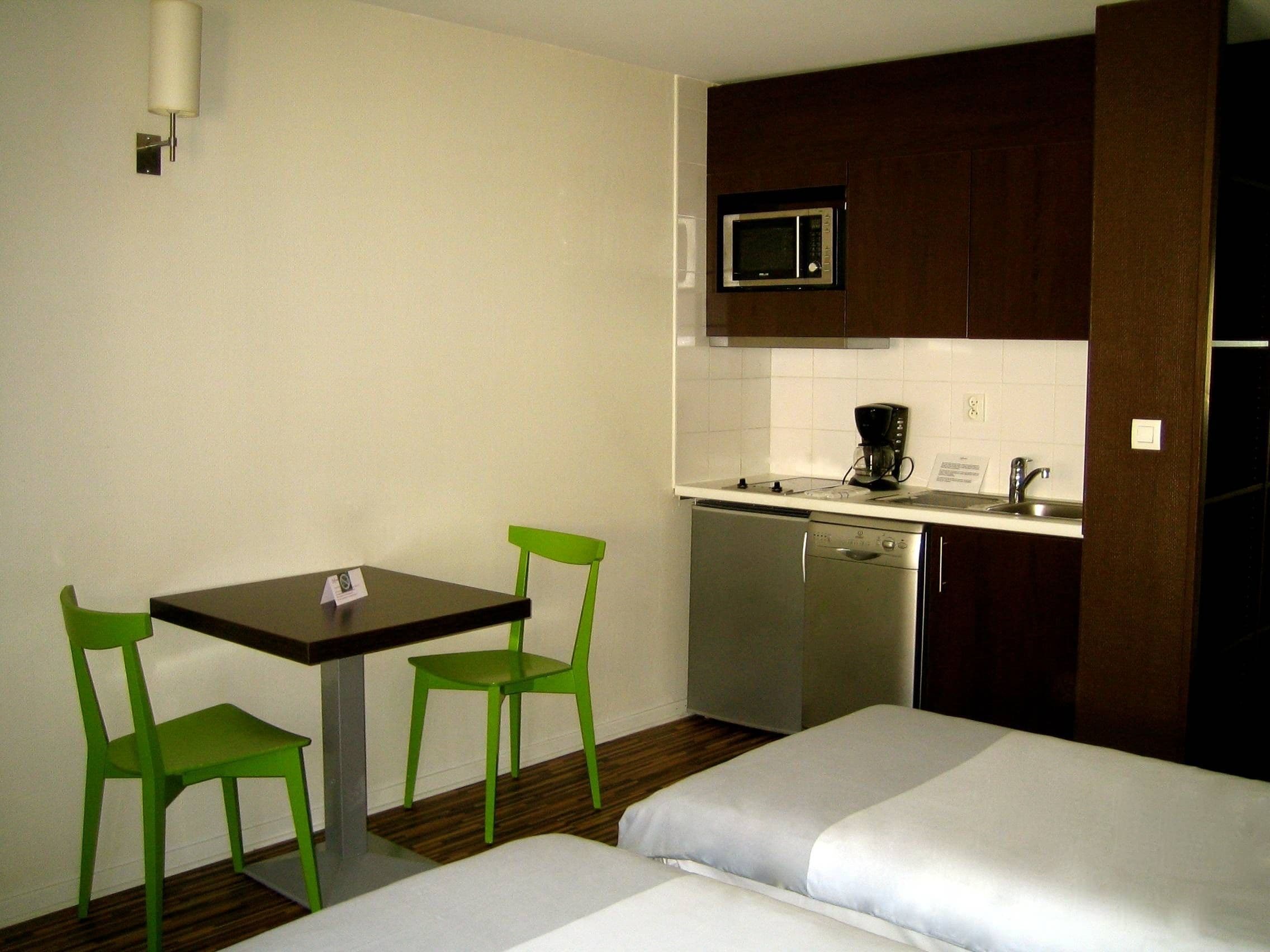 A compact hotel room with a kitchenette, a small dining table with chairs, and two beds.
