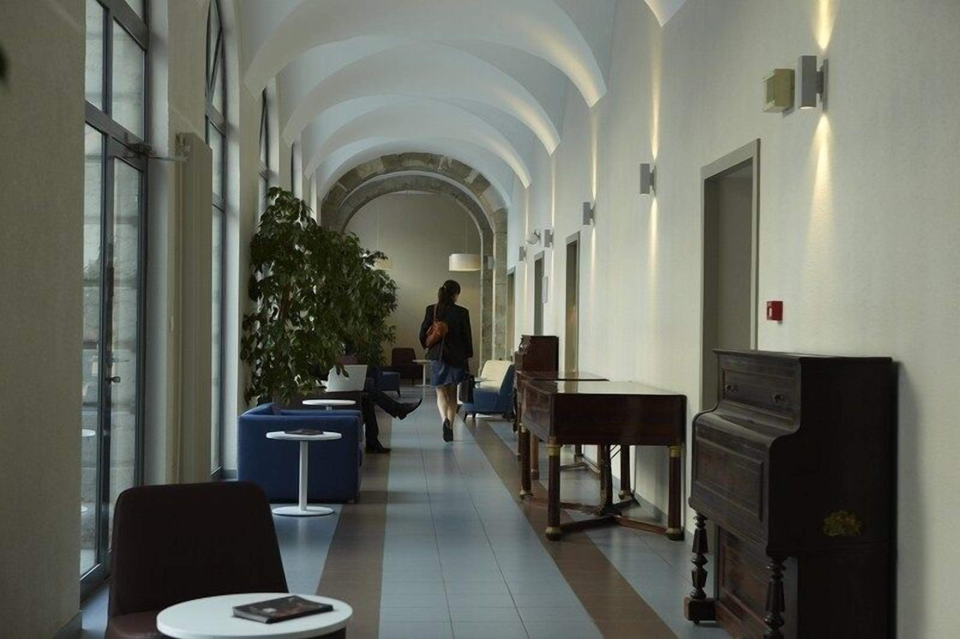 Een hotelgang met een hoge plafond, een grote plant, zithoeken en een piano.