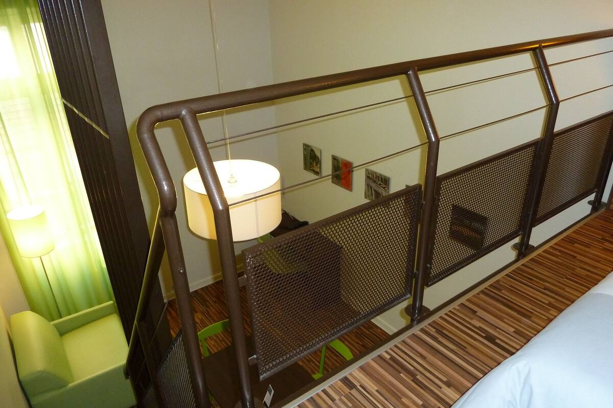 A hotel room with a wooden floor, a bed with white bedding, a green chair, a lamp, and a metal railing.
