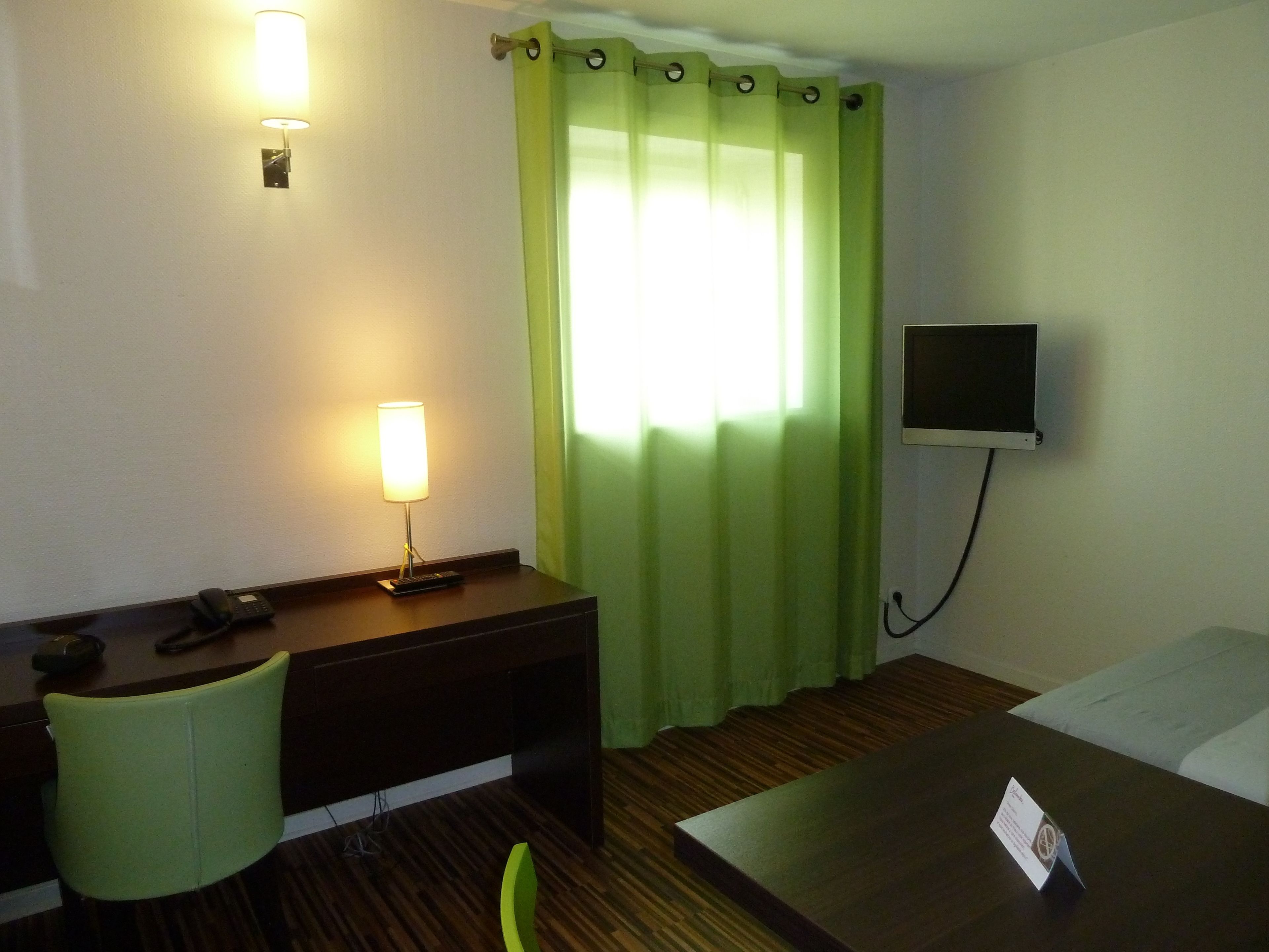 A hotel room with a desk, a chair, a bed, a TV, and a lamp.