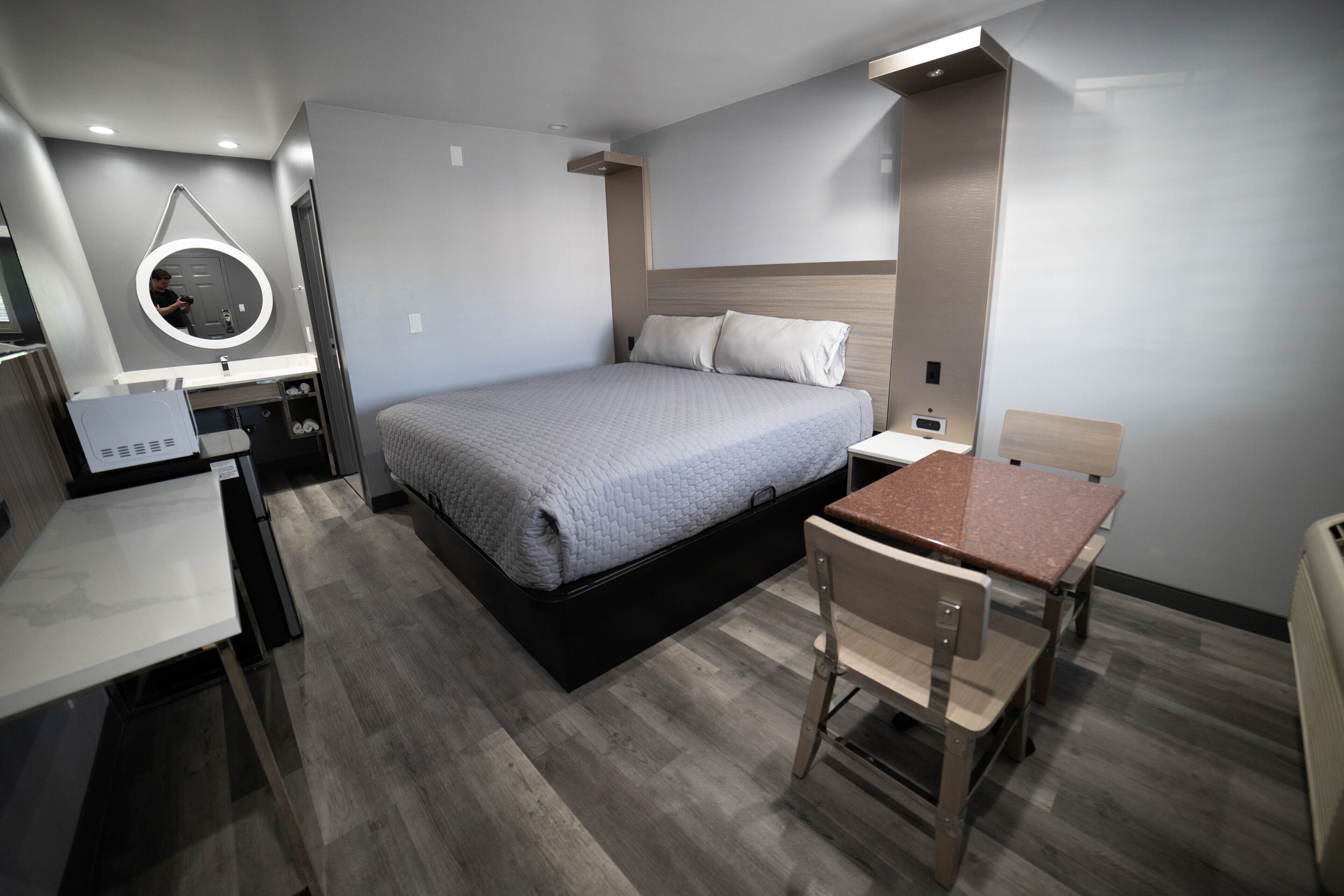A modern hotel room with a bed, a desk, a chair, a round mirror, and a small table.