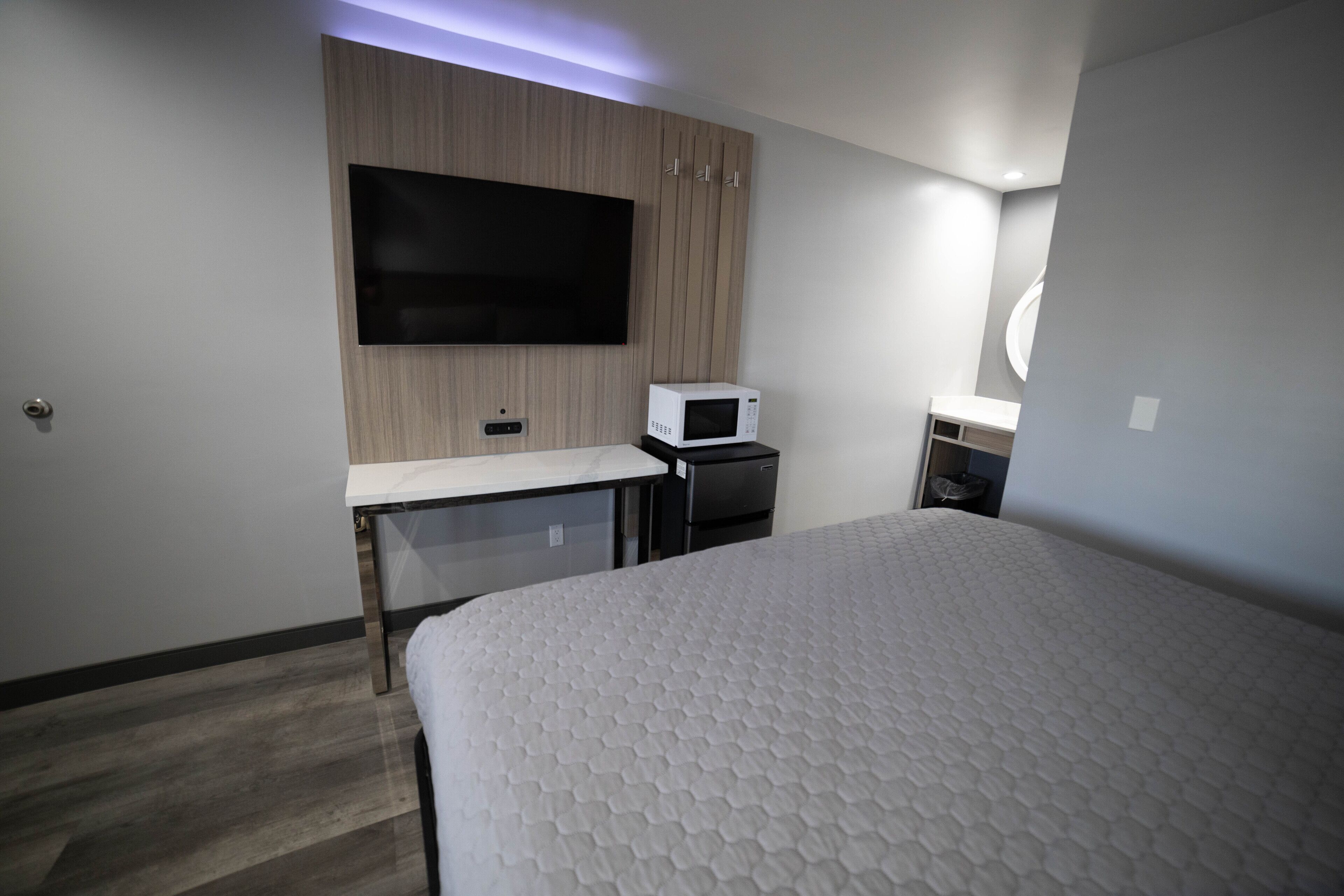 A modern hotel room with a large bed, a flat-screen TV mounted on the wall, a microwave, and a small desk area.