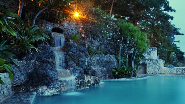 A serene pool area with a waterfall, surrounded by lush greenery and stone structures. - Coralpoint Gardens (Lapu-Lapu)