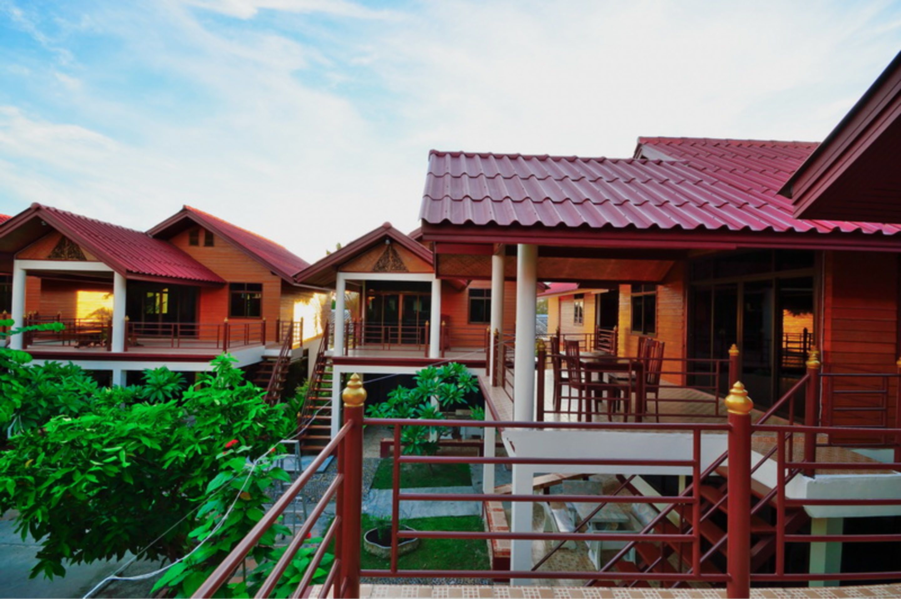 A hotel with multiple wooden buildings, each with a red roof and a balcony. There are stairs connecting the buildings, and the area is surrounded by greenery.