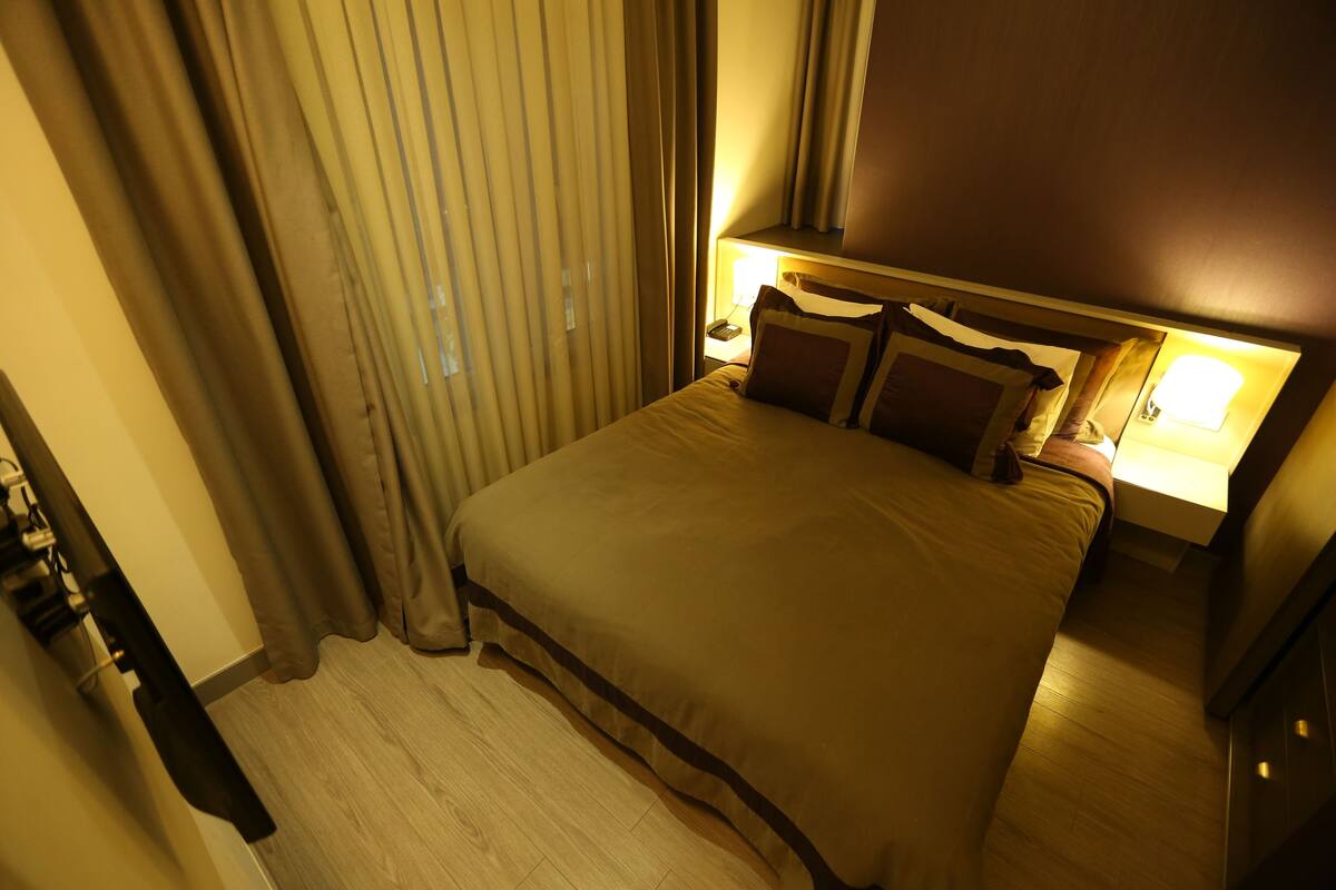 A hotel room with a large bed, a bedside table with a lamp, and a window with curtains.