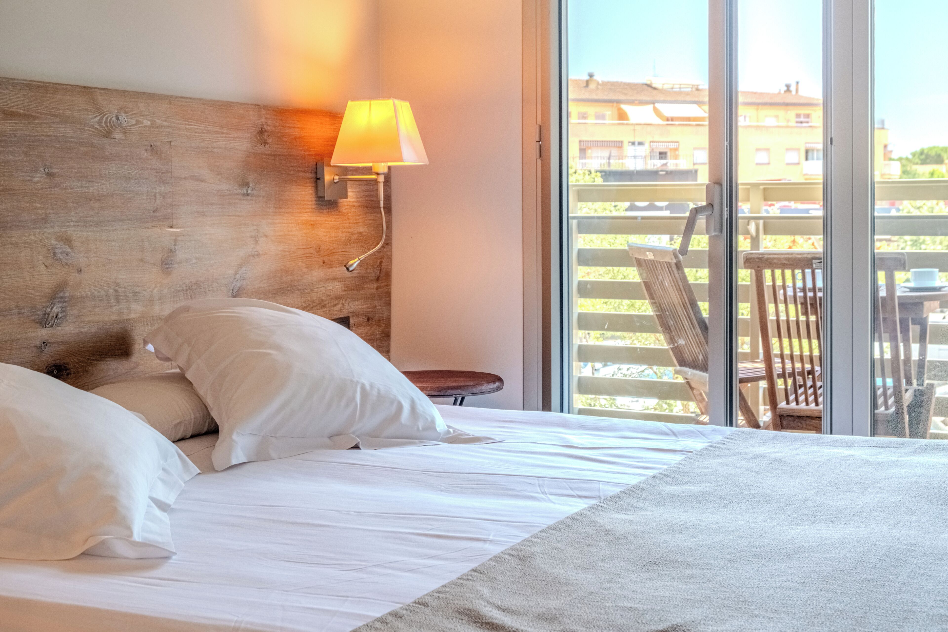 A hotel room with a bed, pillows, a wooden headboard, a wall-mounted lamp, and a view of the outside balcony.