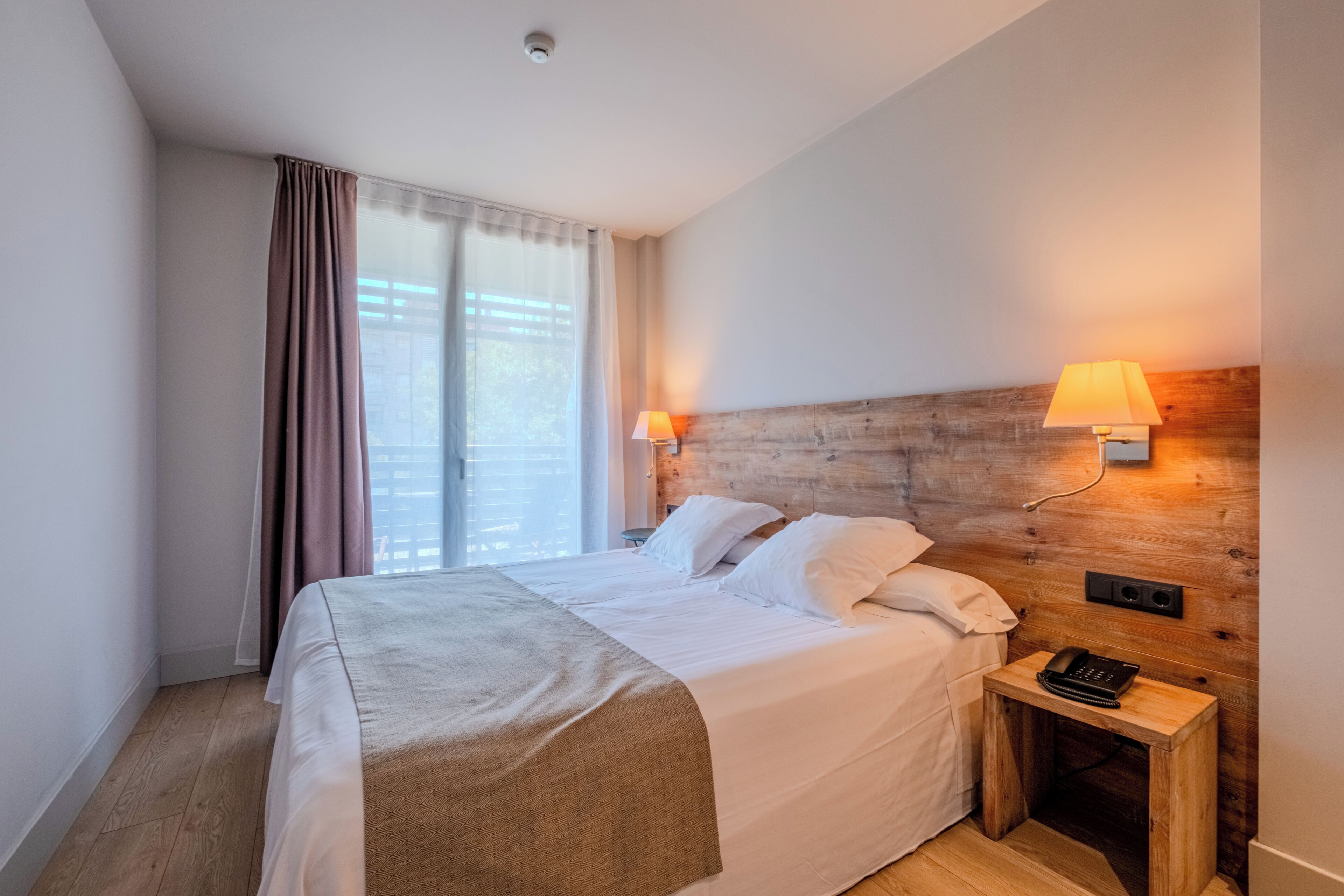 A hotel room with a large bed, two bedside lamps, a wooden headboard, a nightstand with a phone, and a window with curtains.