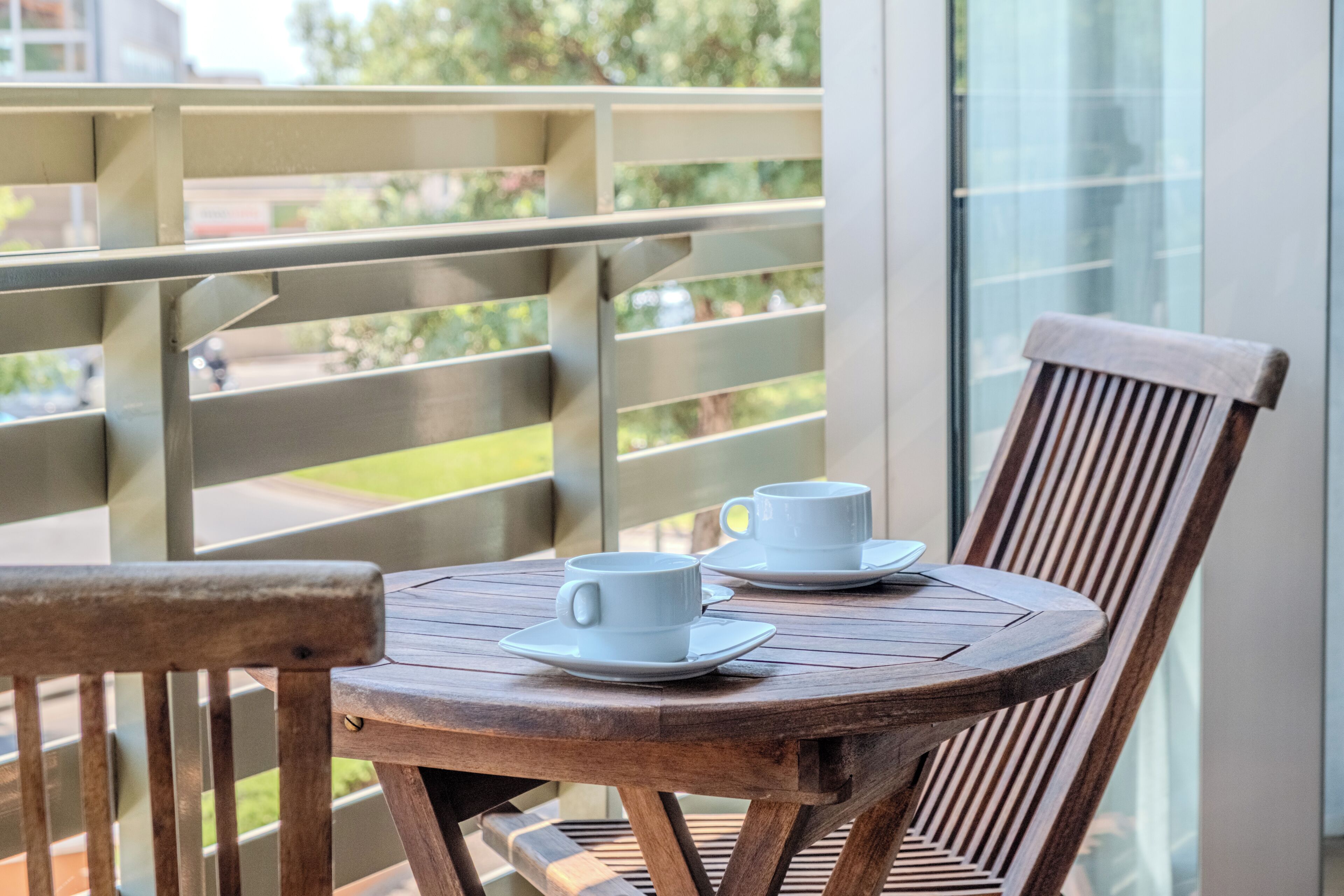 Un balcon avec une table et des chaises en bois, deux tasses blanches et une fenêtre en verre.