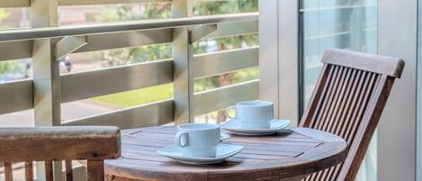 Un balcon avec une table et des chaises en bois, deux tasses blanches et une fenêtre en verre.