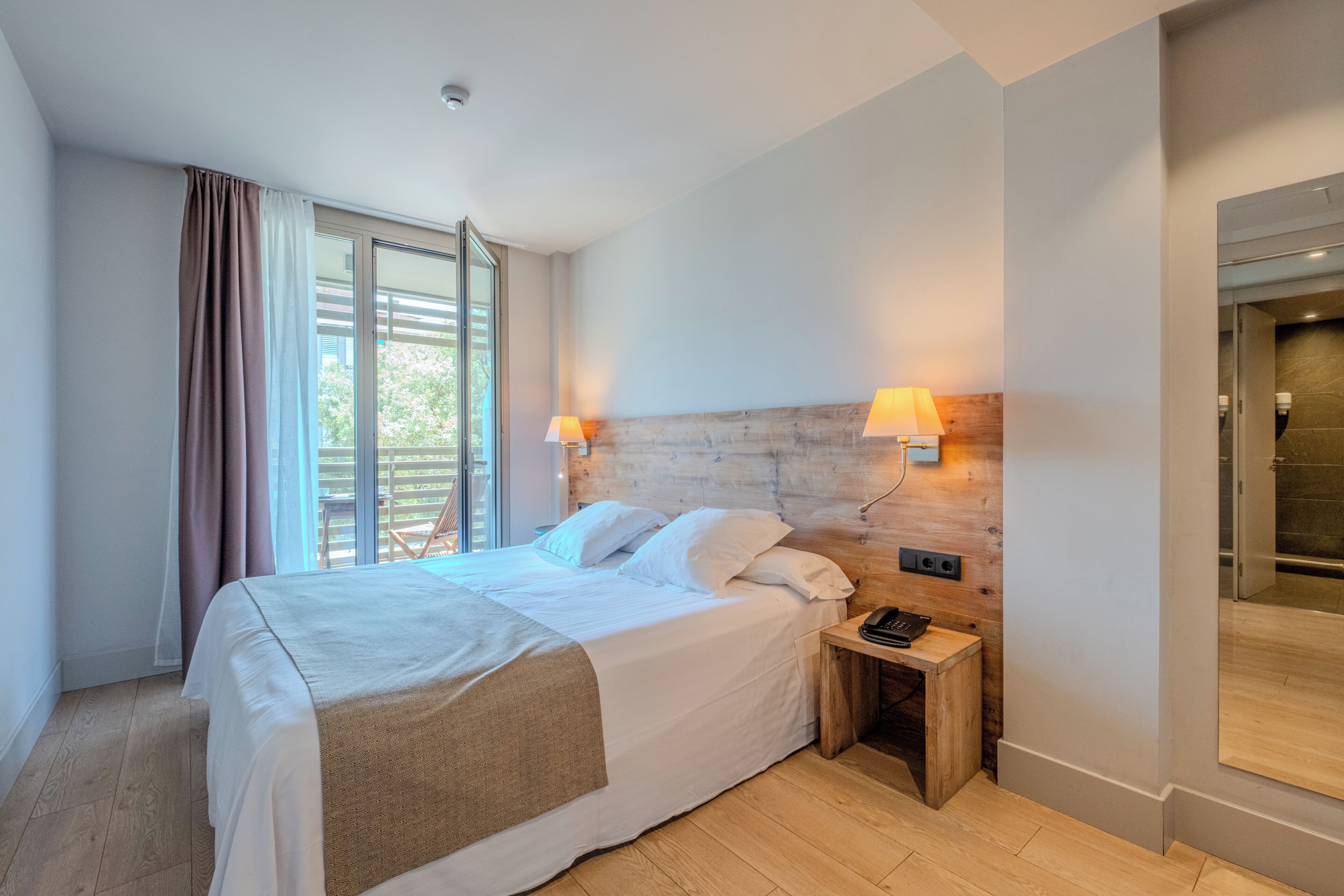 A hotel room with a bed, bedside lamps, a nightstand, and a view of the outdoors through a window.