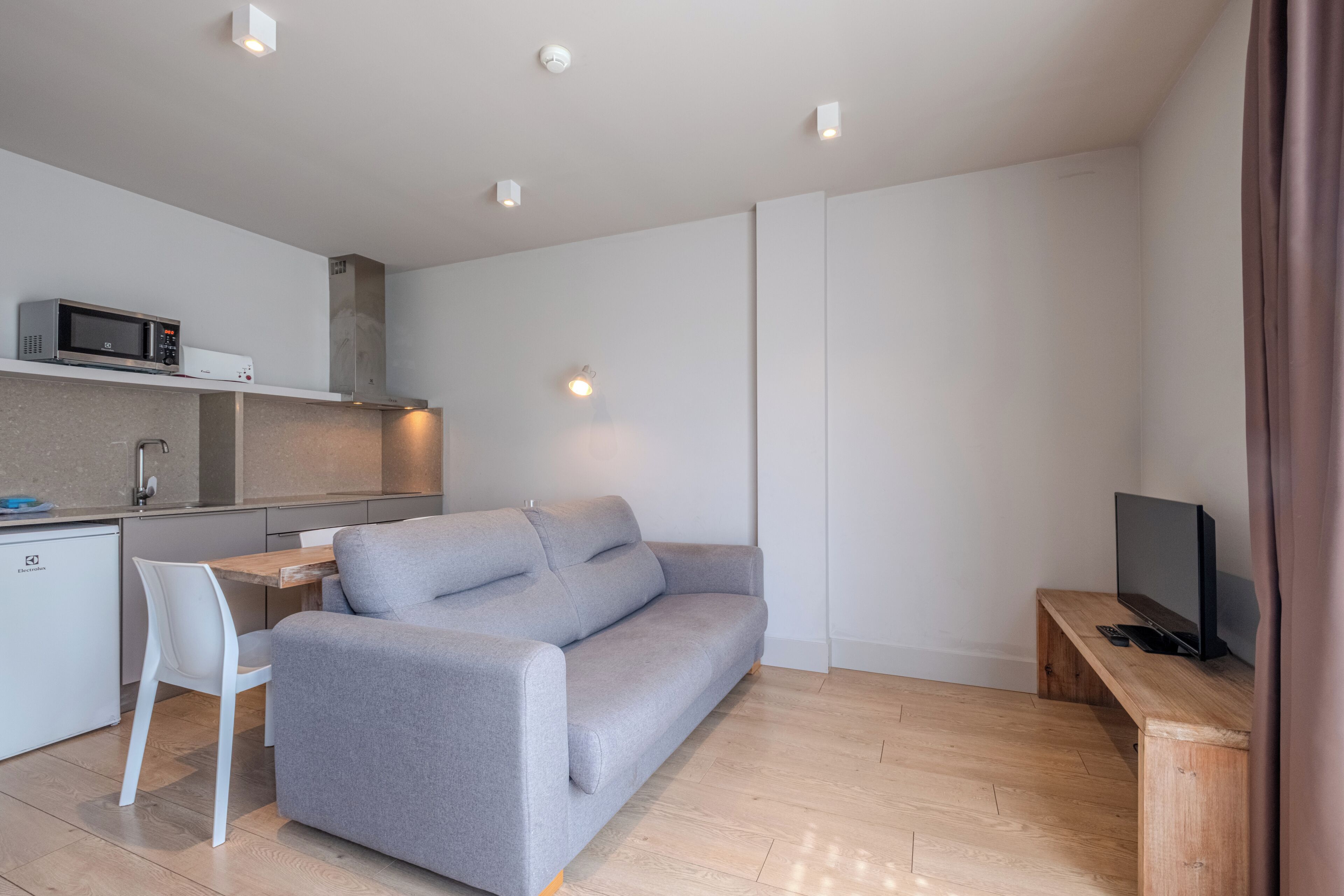 A modern living space with a grey sofa, a wooden TV stand, a white dining table with a chair, a kitchen area with a microwave, and recessed ceiling lighting.