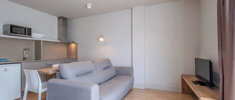 A modern living space with a grey sofa, a wooden TV stand, a white dining table with a chair, a kitchen area with a microwave, and recessed ceiling lighting.