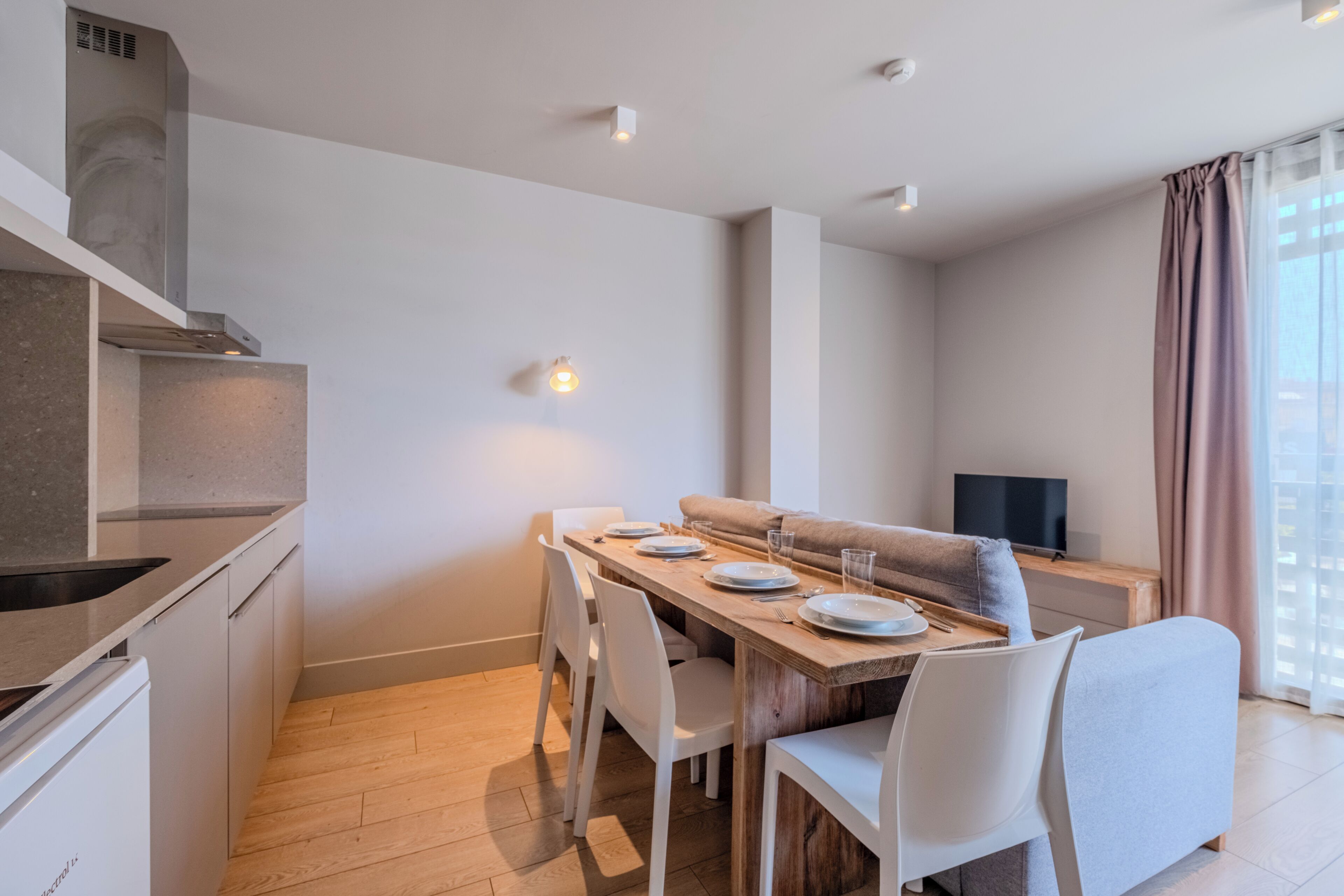 A modern kitchen with a built-in oven, a dining area with a wooden table and white chairs, and a living area with a sofa and a flat-screen TV.