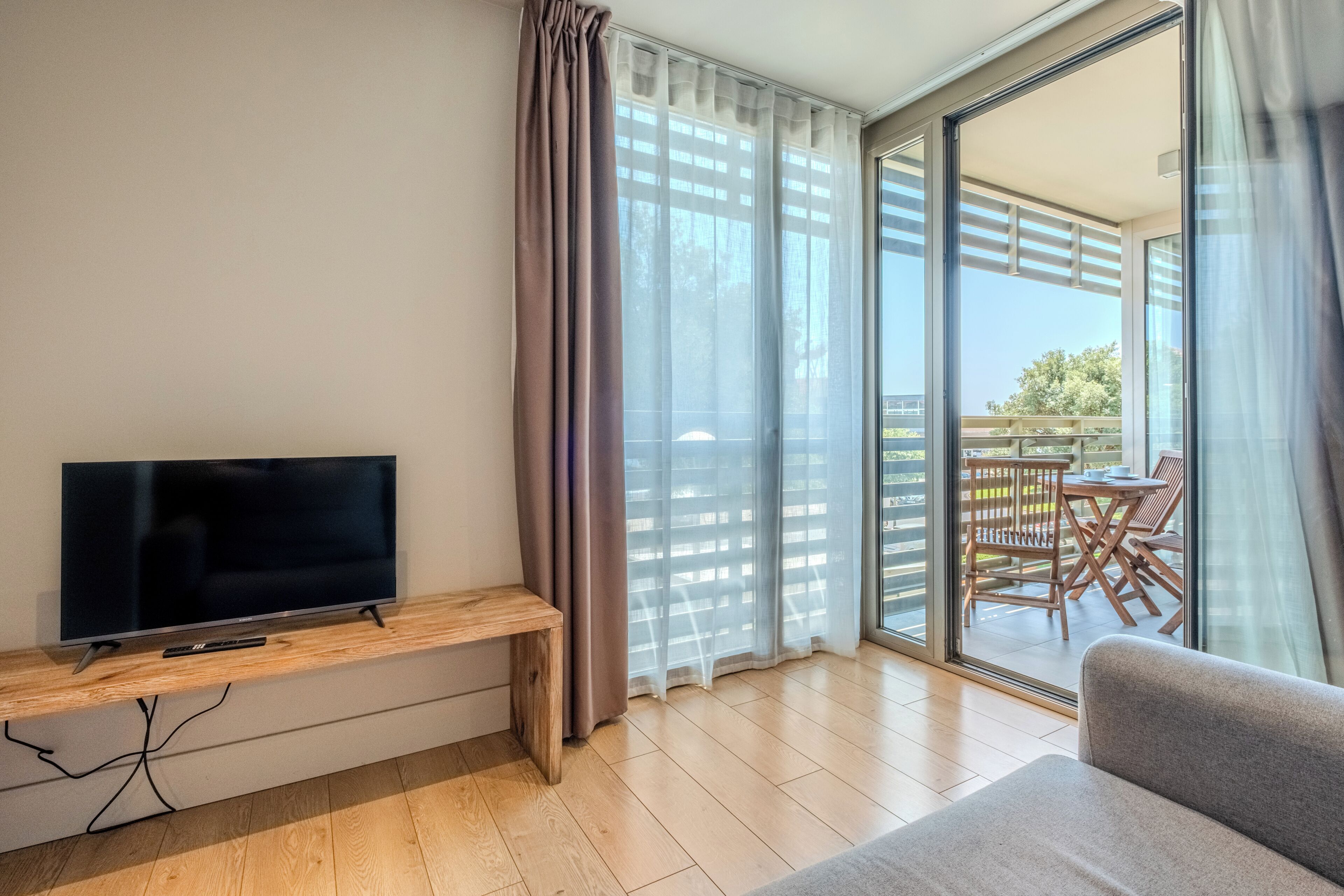 Una sala de estar moderna con una televisión de pantalla plana sobre un mueble de madera, un sofá gris y un ventanal corredizo que da a un balcón exterior con mobiliario de madera.