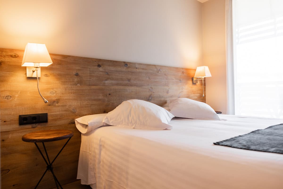 Un lit bien fait, avec du linge de lit blanc, deux lampes fixées au mur, un petit tabouret en bois et une fenêtre avec des stores.