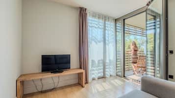 Un saló modern amb un televisor de pantalla plana sobre una cadira de fusta, un sofà gris i una porta corredissa de vidre que dona a una zona exterior.