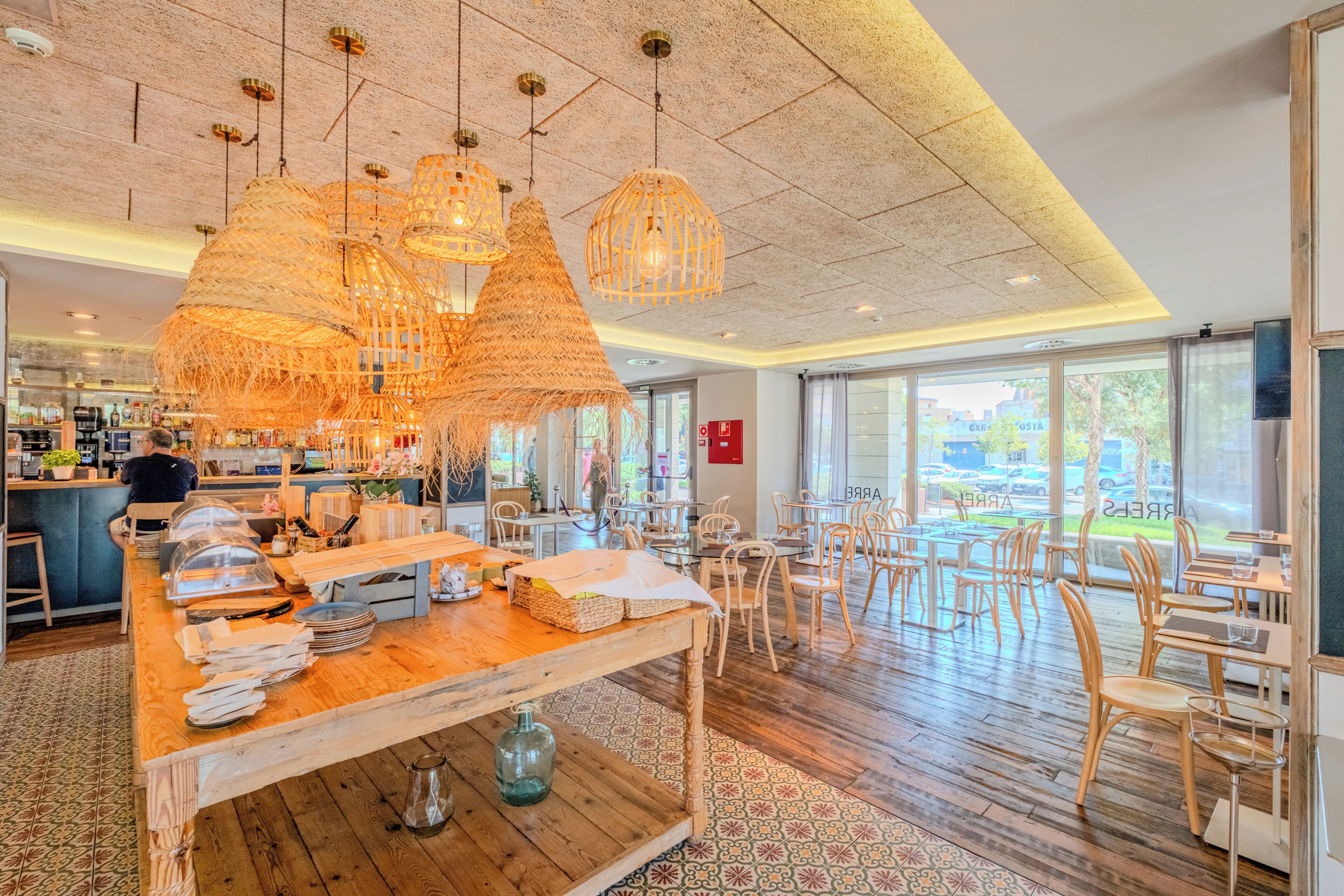 A restaurant interior with a wooden bar area, hanging woven light fixtures, and a view through large windows.