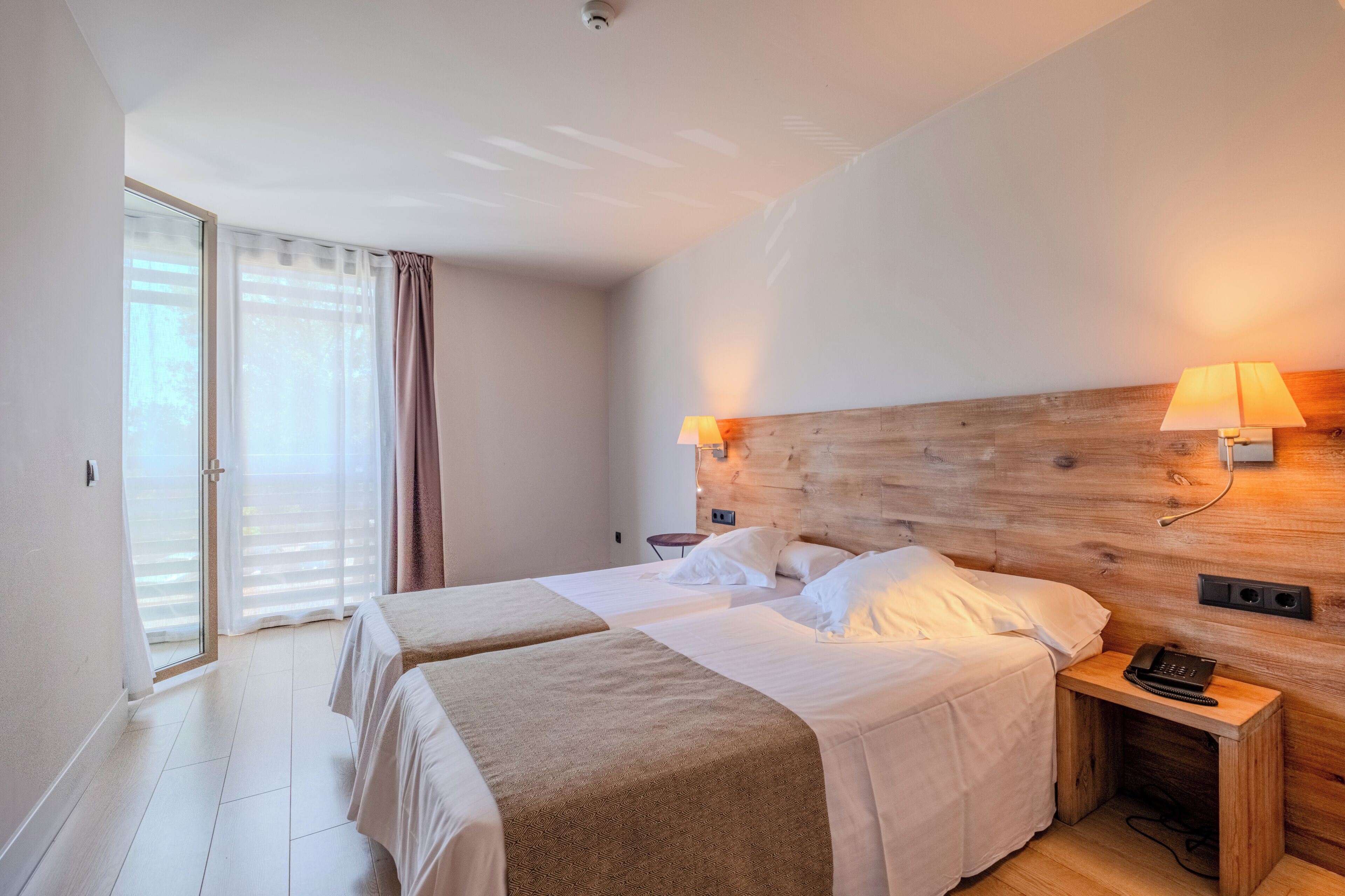Una habitación de hotel con cabecera de madera, dos camas, una mesita de noche con lámpara y un teléfono.