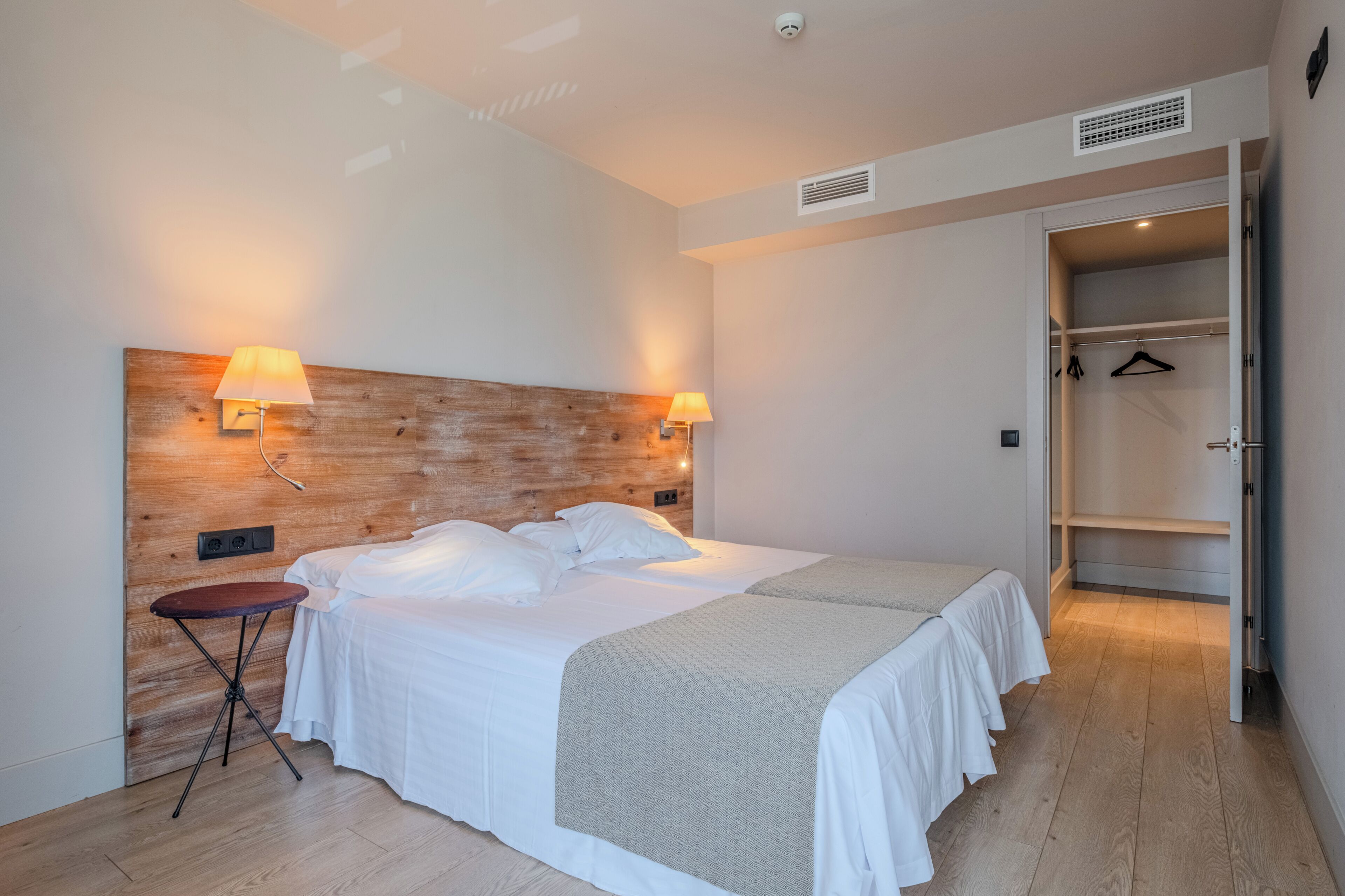 Un dormitorio con cabecera de madera, dos lámparas de noche, una cama con ropa de cama blanca, una mesita y un vestidor.