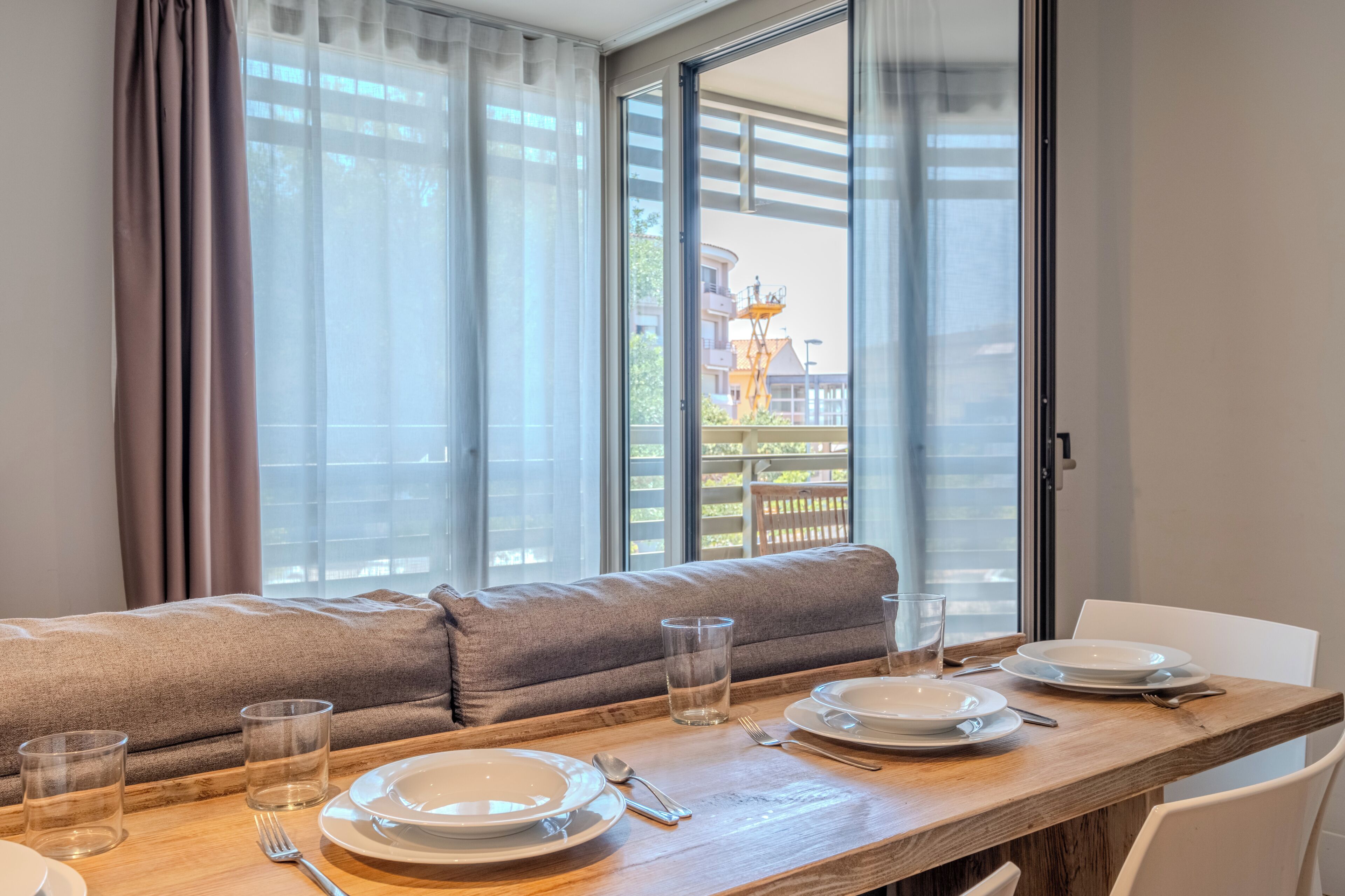 Un comedor con una mesa de madera dispuesta para dos, un sofá y vistas a un edificio con una grúa.