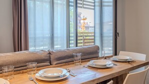 Un coin repas avec une table en bois dressée pour deux personnes, un canapé et une vue sur un immeuble avec une grue à l’extérieur.