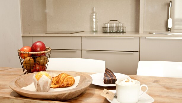 Une table en bois dressée pour le petit-déjeuner, avec un panier de fruits, un croissant, un gâteau au chocolat et une tasse de café.