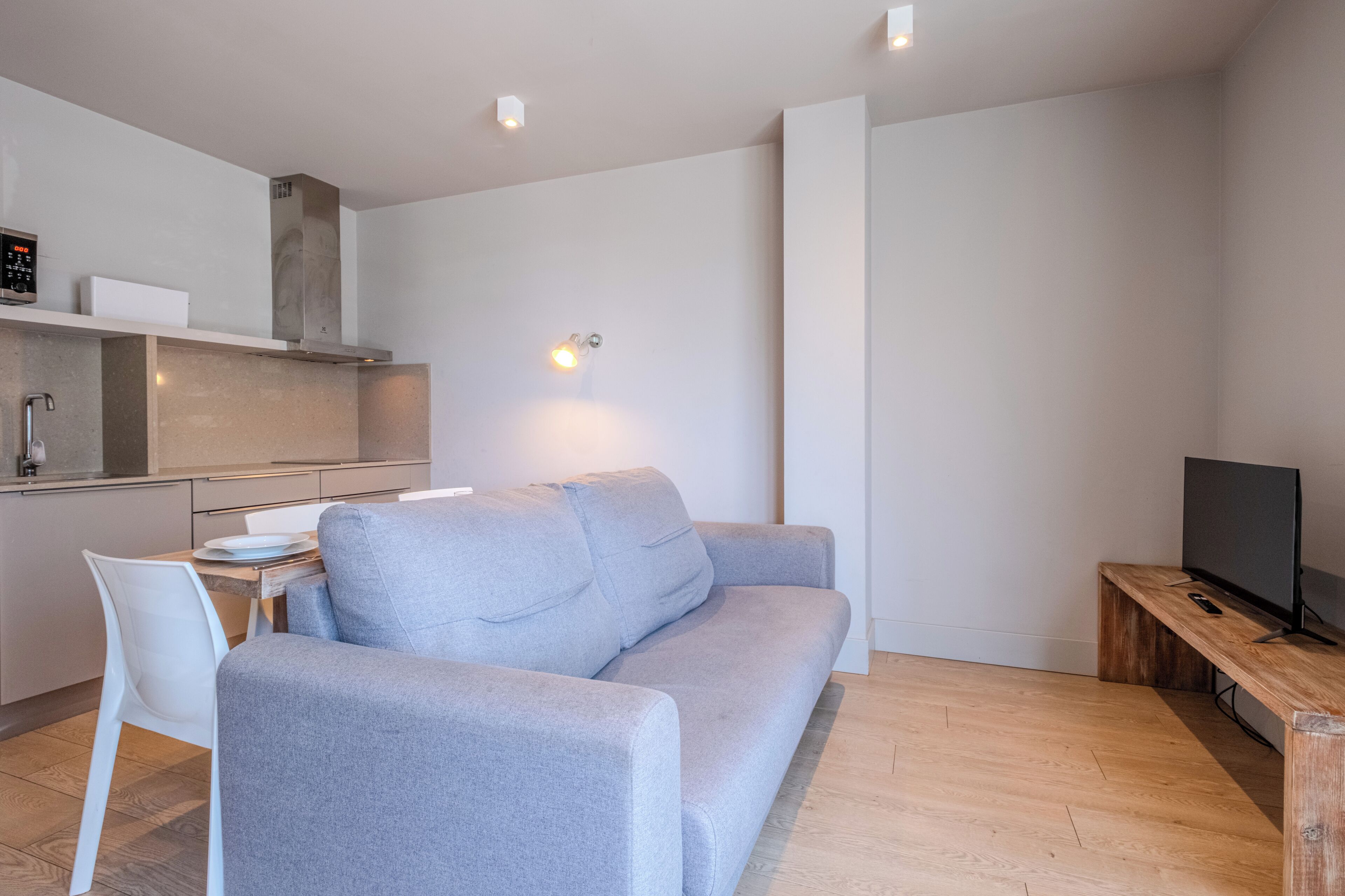 Ein modernes Wohnzimmer mit grauem Sofa, einem Holztisch, einem Fernseher und einer Küchenzeile mit Herd und Schränken.