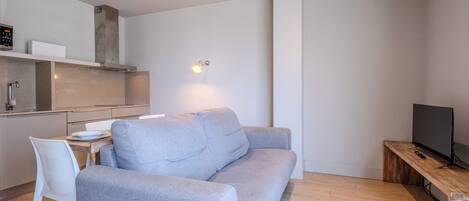 Een moderne woonruimte met een grijze bank, een houten salontafel, een televisie en een keuken met een fornuis en kastjes.