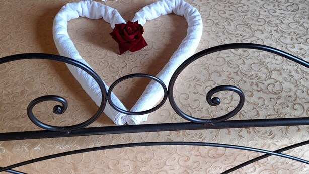 Ein Bett mit dekorativer Handtuchgestaltung in Form von Schwänen und einer Rose, einem roten Kissen und einer gemusterten Bettdecke.