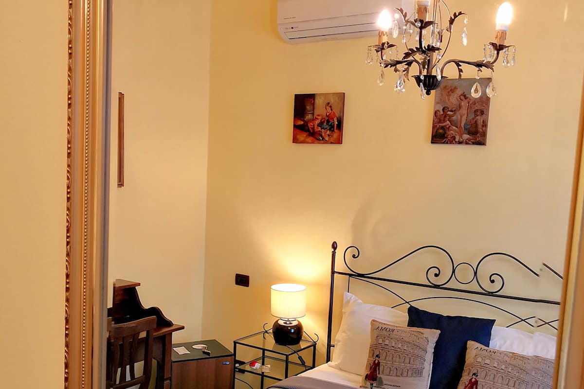 A bedroom with a bed, a chandelier, an air conditioner, a desk, and a chair.