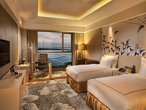 Ein Hotelzimmer mit zwei Betten, einem Schreibtisch, einem Fernseher, einem Fenster mit Blick auf den Strand und einem gemusterten Teppich.