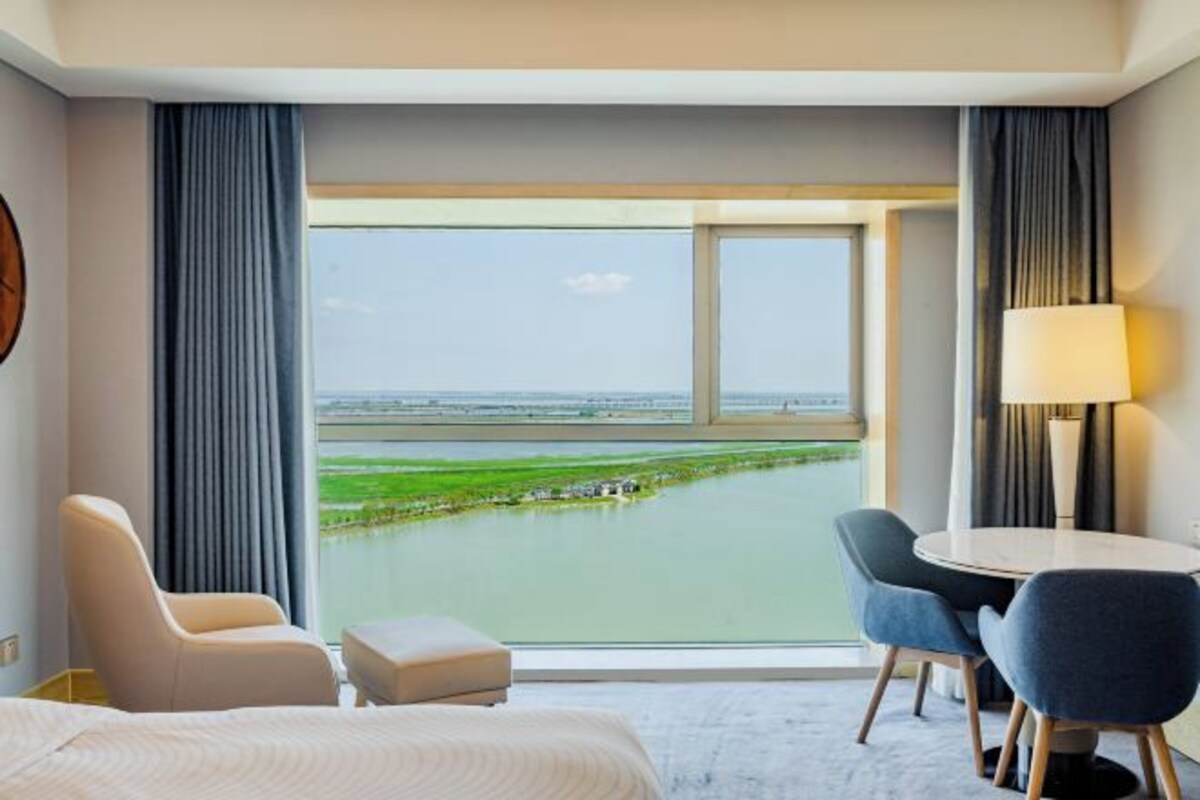 A hotel room with a large window offering a view of a body of water and greenery. There is a bed, a chair, and a small table with a lamp.