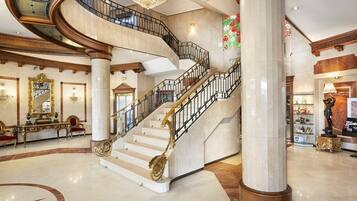 A grand lobby with a marble floor, a curved staircase with golden railings, and a large chandelier.