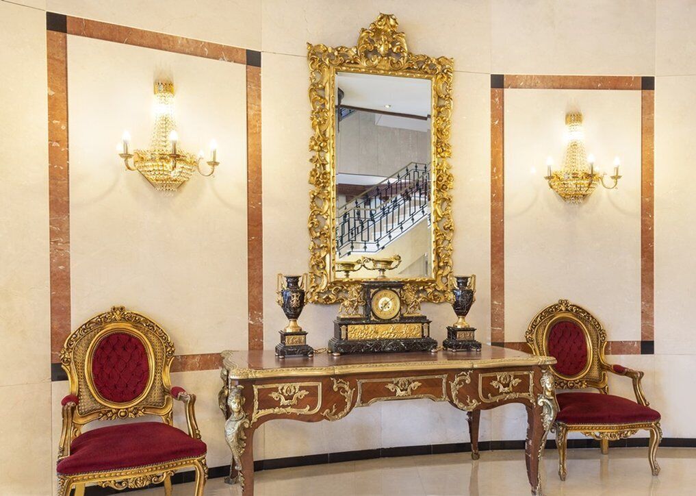 A luxurious room with a large ornate mirror, two gold-framed chairs, and a matching table with decorative items.