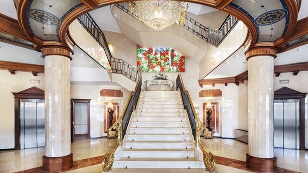 Una imponente escalera con escalones de mármol y barandillas doradas, flanqueada por columnas monumentales y un candelabro.