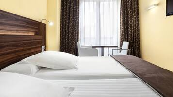Habitación de hotel con cabecera de madera, cama con ropa blanca, una mesita con una silla y una ventana con cortinas.
