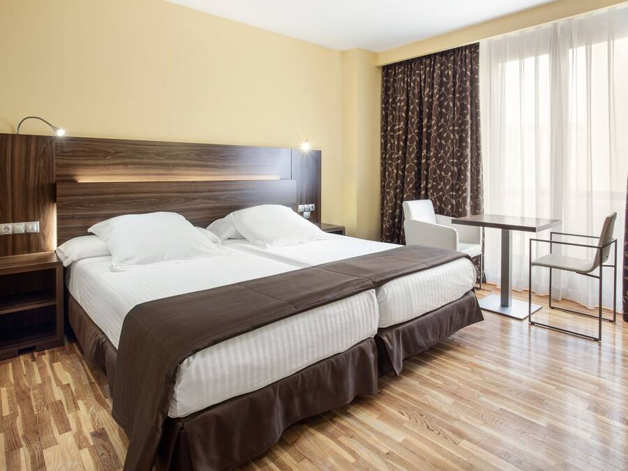 Een hotelkamer met een groot bed, een nachtkastje, een bureau met stoel en een raam met gordijnen.